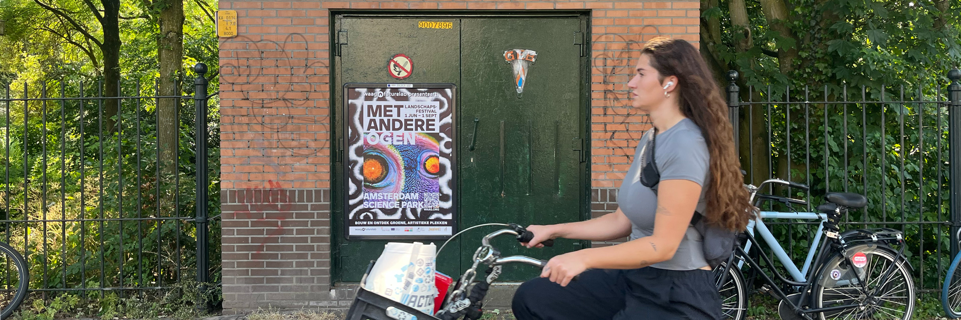 Photograph of a woman on a bicycle passing a poster on a wall. The poster reads ‘Met Andere Ogen’.